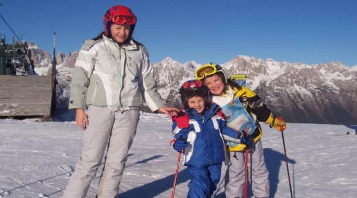 Tre ragazzini tra cui due ragazze ed un bambino posano sorridenti con i bastoni da sci in mano. Ai loro piedi si stende un manto di neve e sullo sfondo si intravedono delle vette innevate.