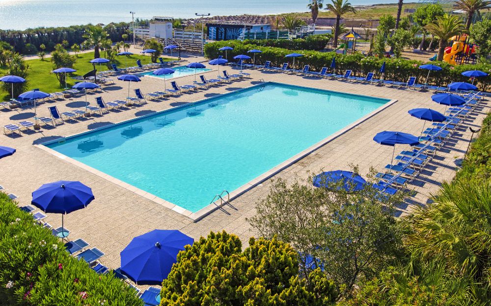 Hotel African Beach. Una piscina rettangolare dall'acqua cristallina si estende al centro della composizione, circondata da lettini e ombrelloni blu. Ai lati si intravede del prato verde e ben curato.