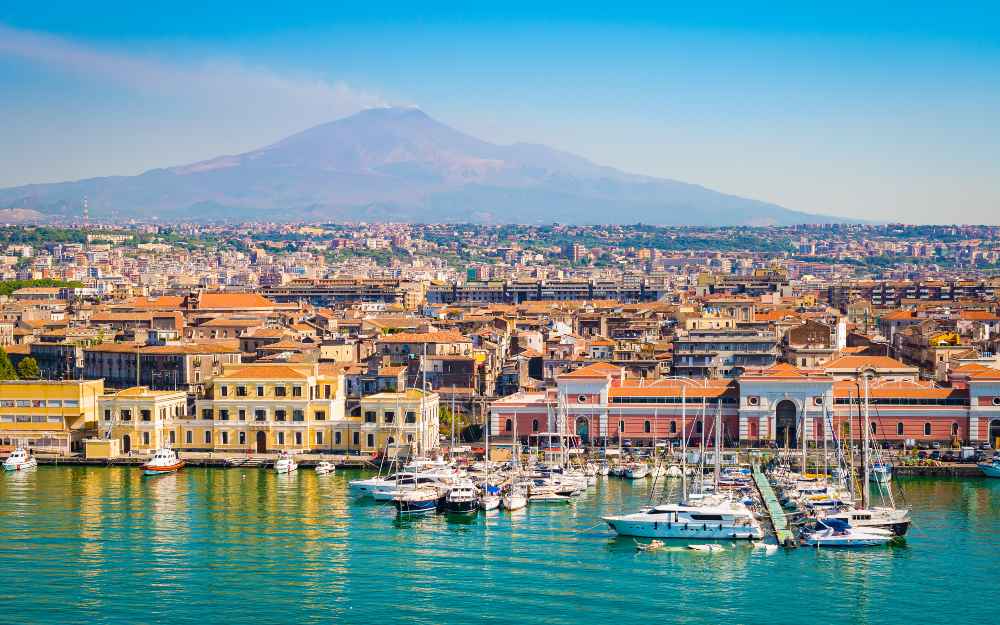 la acque turchesi ospitano qualche imbarcazione mentre sulla sponda del porto si intravedono delle facciate di case colorate, incorniciate ina alto dal profilo del Vesuvio.