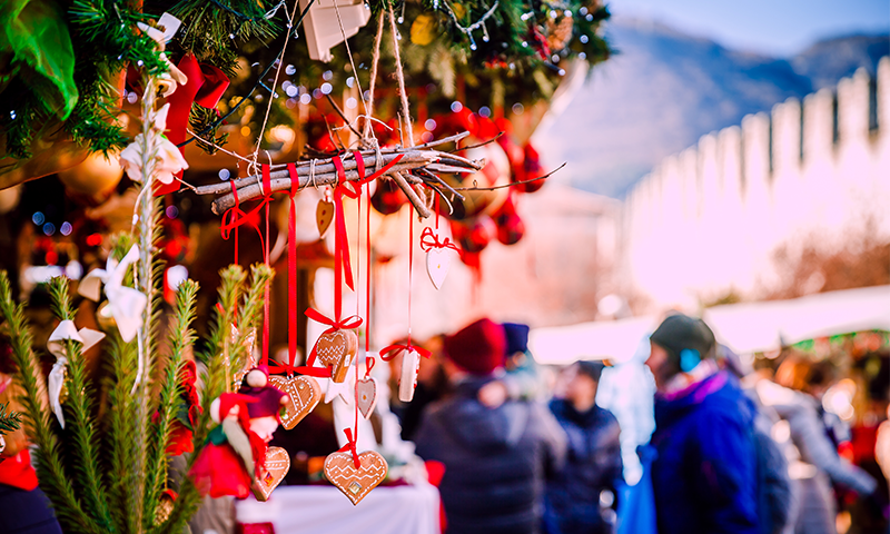 Mercatini di Natale di Trento, in piazza Fiera. Sulla sinistra in primo piano si notano i decori Natalizi della casetta, mentre sulla destra passeggiano i passanti, con le mura medievali della piazza che fanno da sfondo.