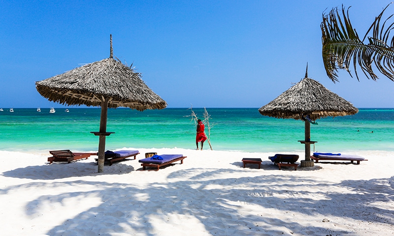 Un kenyota in abiti tradizionali sistema una rete su una spiaggia di Watamu, di sabbia bianca e fina, con un paio di ombrelloni di paglia. Sullo sfondo il mare turchese che abbraccia la spiaggia ed il cielo limpido.